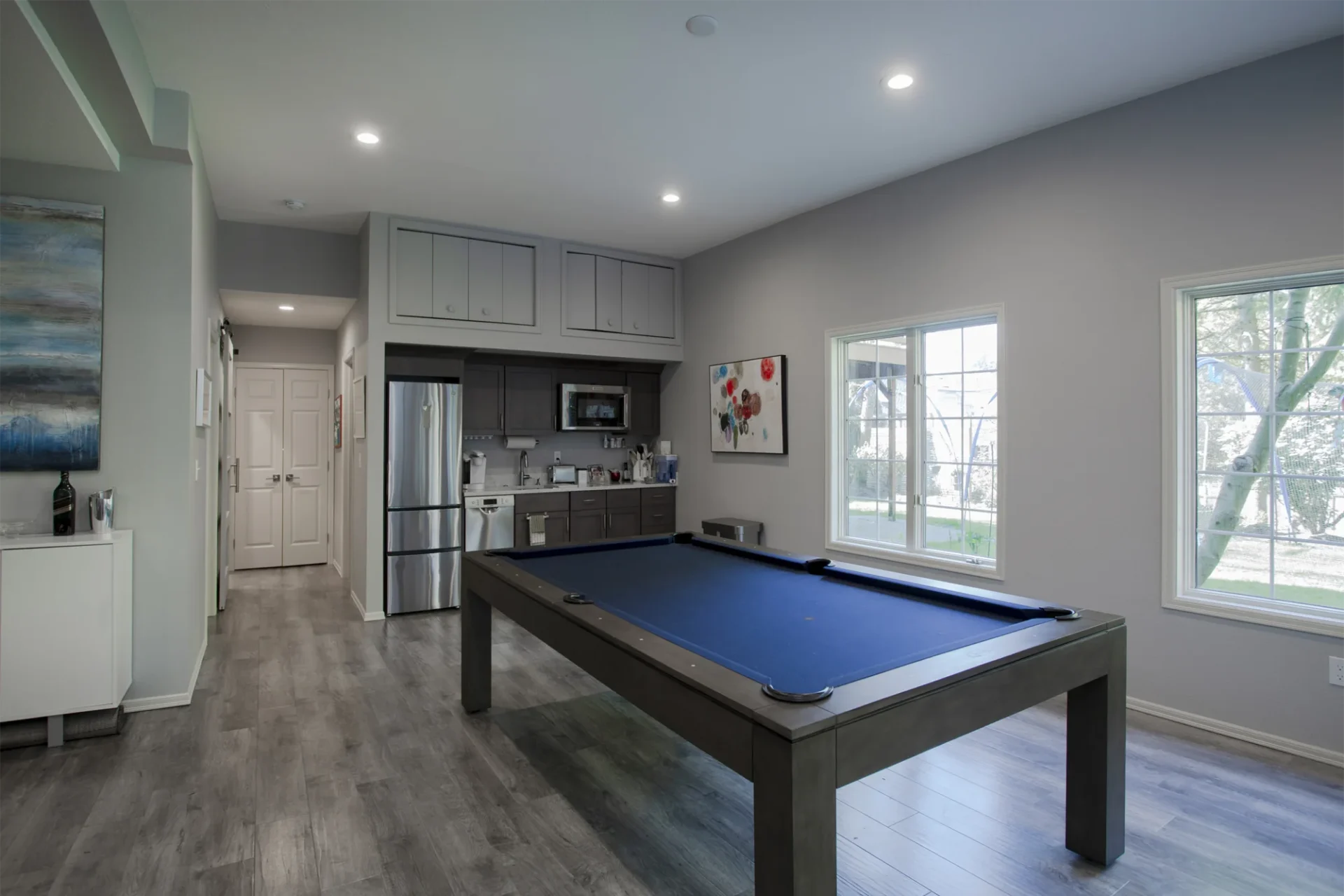 Modern game room remodel by Home Run Design Remodel with blue pool table, kitchenette, stainless appliances, and large windows.